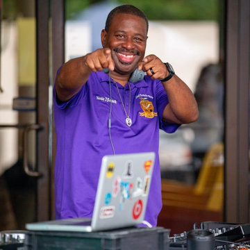 Man in a purple shirt with a logo, standing behind DJ equipment outdoors.